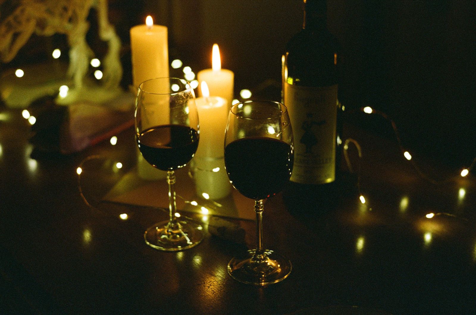 Two glasses of red wine beside a bottle, candles and string lights
