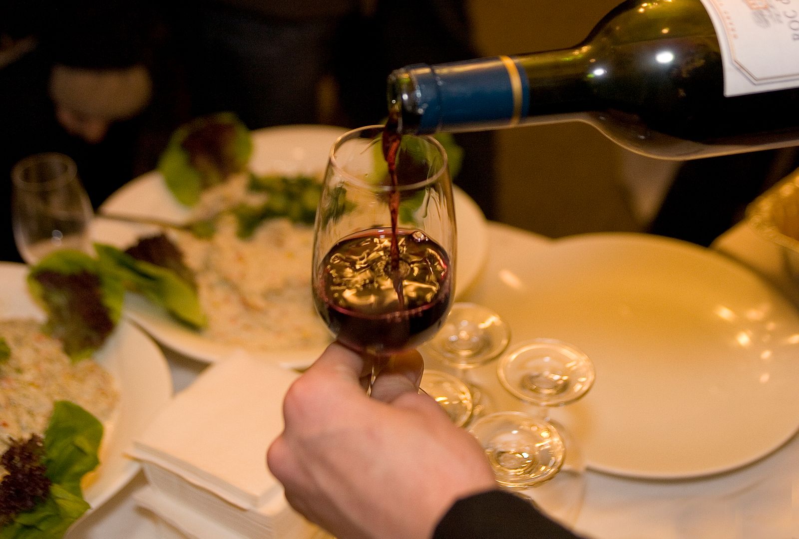 A bottle of red wine being poured into a glass at a table of shared plates