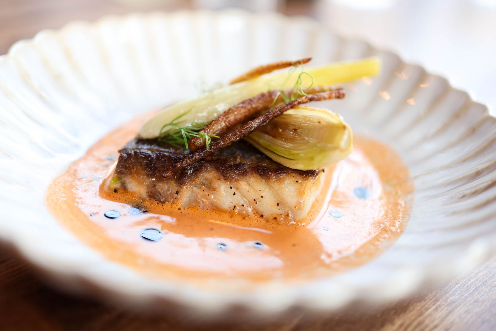 A seared fish fillet in an orange sauce on a scalloped white plate